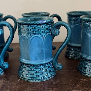 May include: A set of five ceramic mugs in a deep teal blue glaze. Each mug features a textured basket-weave pattern at the base and rim, with a smooth arched panel design in the centre and a curved handle extending from the side of the vessel.