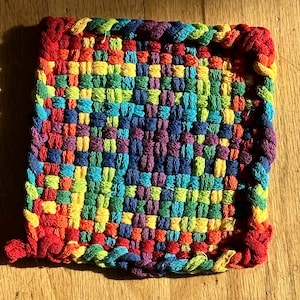 Könnte beinhalten: Ein quadratischer, handgewebter Topflappen in einem lebendigen Regenbogen aus Farben. Das gewebte Design weist ein sich wiederholendes Muster aus Rot, Orange, Gelb, Grün, Blau und Lila auf. Die Kanten sind mit einem geflochtenen Rand in passenden Farben versehen.