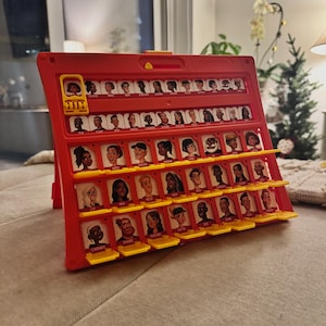 Può includere: Un gioco da tavolo Guess Who? rosso con pannelli scorrevoli gialli. Il gioco presenta piccoli ritratti di varie persone. Il gioco è progettato per essere giocato da due giocatori, che cercano di indovinare il personaggio misterioso dell'altro giocatore ponendo domande sì o no.