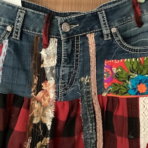 May include: A denim skirt with patchwork details. The skirt features a combination of blue denim, red and black plaid fabric, floral prints, and lace accents. Decorative stitching and a button closure are visible.