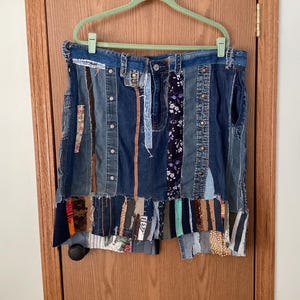 May include: A denim skirt with a patchwork design. The skirt is made from various shades of blue denim, with strips of fabric in different patterns and colors sewn together. It hangs on a green hanger against a wooden door.