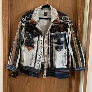 May include: A unique, light-wash denim jacket with intricate patchwork and embellishments. Features include decorative stitching, lace, and various fabric strips in shades of blue, brown, and red. The jacket is hanging on a wooden door.