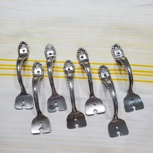 May include: Eight silver-toned fork-shaped drawer pulls with ornate details. The pulls have a decorative design at the top and a cut-out shape at the bottom.