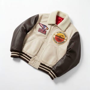 May include: Cream-colored bomber jacket with brown leather sleeves and a cream-colored shearling collar. Features embroidered patches with "USA" and "Flying Legends American Airborne" text. The jacket has a striped brown and cream ribbed hem and cuffs.