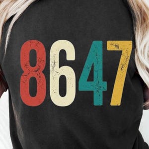 Peut inclure: T-shirt noir avec le numéro "8647" imprimé dans un style rétro. Les chiffres sont rouges, crème, turquoise et jaunes. Le t-shirt est à col rond.