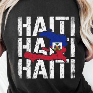 Peut inclure: T-shirt noir avec le mot "HAITI" en blanc, répété trois fois dans une police vieillie. Le drapeau haïtien est affiché au centre, avec un contour de carte rouge et bleu. Le t-shirt est à col rond.
