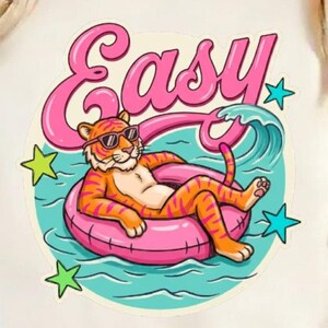 Peut inclure: T-shirt crème avec un tigre de dessin animé portant des lunettes de soleil, se relaxant sur un anneau gonflable rose dans l'eau. Le mot "Easy" est en script rose au-dessus du tigre. Des étoiles vertes et bleues ornent l'image.