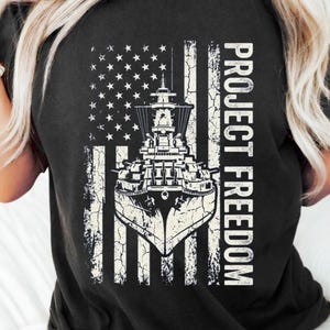 Peut inclure: T-shirt noir avec un drapeau américain vieilli, un dessin de cuirassé et le texte "PROJECT FREEDOM" en blanc. Le t-shirt a un col rond et des manches courtes.