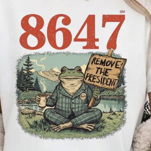 Peut inclure: T-shirt blanc avec un dessin d'une grenouille en costume à carreaux tenant une pancarte « REMOVE THE PRESIDENT ». Le chiffre « 8647 » est imprimé au-dessus de la grenouille. La grenouille est assise devant un lac et des montagnes.