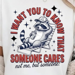 Peut inclure: T-shirt blanc avec un dessin d'un raton laveur dans une chaise longue, portant une casquette et buvant dans une tasse. Le texte dit "I WANT YOU TO KNOW THAT SOMEONE CARES, not me, but someone !" Étoiles noires et texte rouge.