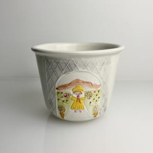 May include: A white ceramic cup featuring a raised, textured diamond pattern on the exterior. A central embossed scene depicts a figure in a yellow dress standing under a brown archway, surrounded by stylized trees with small red and green dots.
