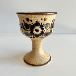 May include: A ceramic goblet with a beige speckled finish, adorned with a band of blue floral designs. The goblet has a flared rim and a pedestal base, with brown accents. The overall design is rustic and decorative.