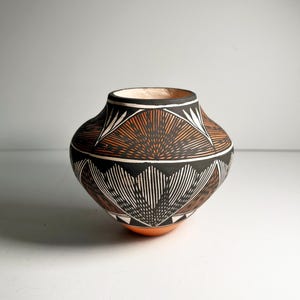 May include: A handcrafted pottery vessel with a rounded body and a wide opening. The pot features intricate geometric designs in black, white, and orange. The base is orange, and the interior is a light peach color. The vessel is likely used for decorative purposes.