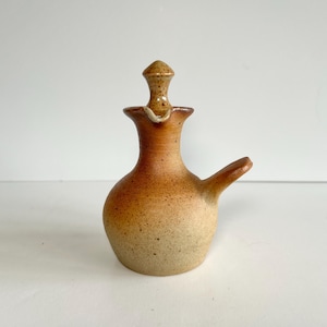 Puede incluir: Una pequeña jarra de cerámica hecha a mano, con un pico y un tapón. La jarra tiene una base redondeada y un cuello delgado, con un degradado de tonos marrones y beige. El tapón es del mismo color y forma. La jarra es probablemente para servir líquidos.