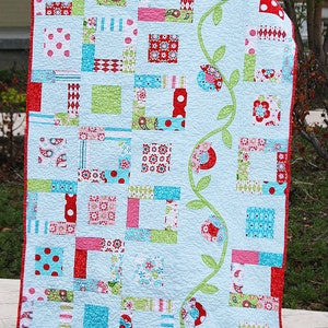 May include: A blue and white quilt with a green vine and red flowers along the side. The quilt is made up of squares of different fabrics with various patterns, including polka dots, stripes, and floral prints. The quilt is labeled "Sew Charming" in the bottom right corner.