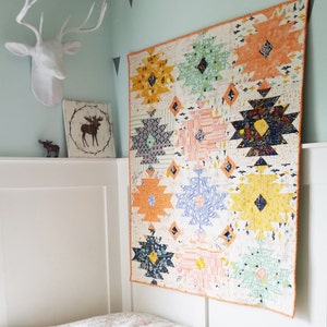 Puede incluir: Una colcha colorida con un patr&oacute;n geom&eacute;trico en tonos de naranja, amarillo, azul, verde y blanco. La colcha est&aacute; colgada en una pared blanca en una habitaci&oacute;n con un techo blanco y paredes de color azul claro.