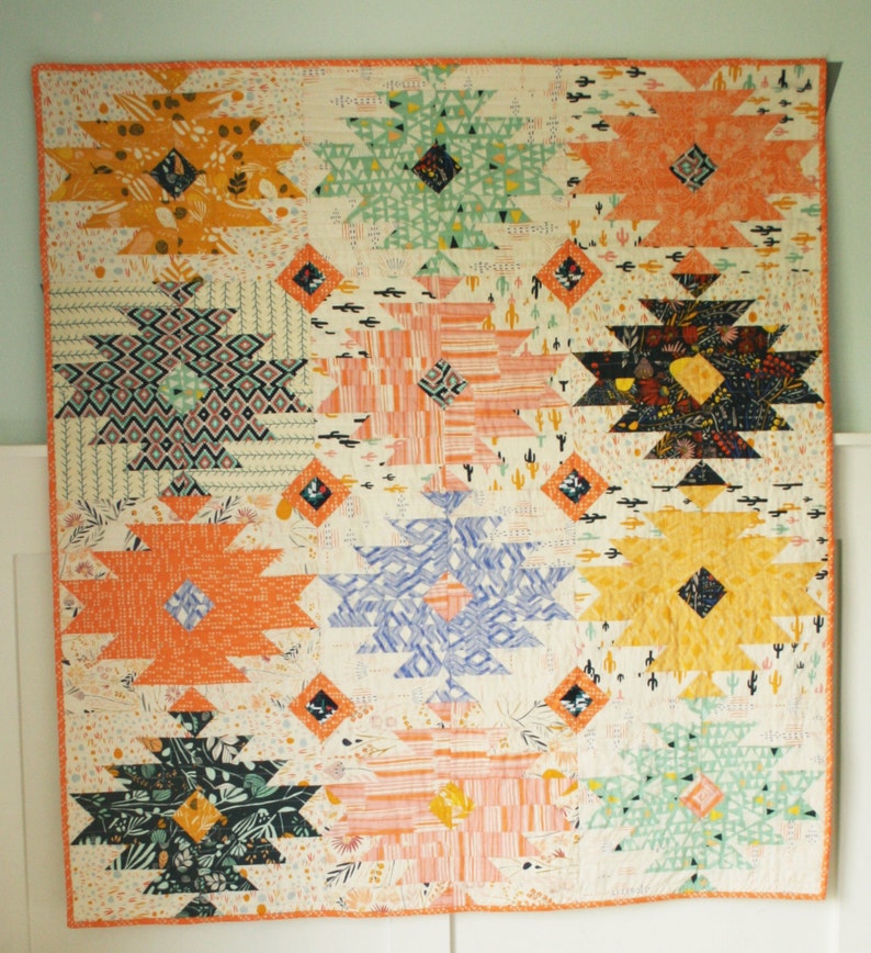 Puede incluir: Una colcha colorida con un patr&oacute;n geom&eacute;trico de estrellas y diamantes. La colcha est&aacute; hecha de varias telas en tonos de naranja, amarillo, verde, azul y negro. La tela de fondo es blanca con un peque&ntilde;o estampado de cactus.