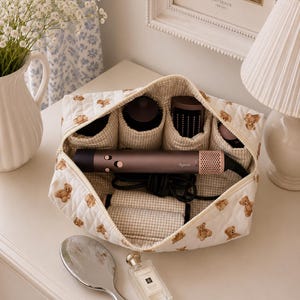 Op de afbeelding: Een witte, gewatteerde cosmetische tas met een teddybeerpatroon, open om haarstylingtools te onthullen. Een zilveren handspiegel en een parfumflesje liggen op het witte oppervlak. Een Dyson föhn is zichtbaar.