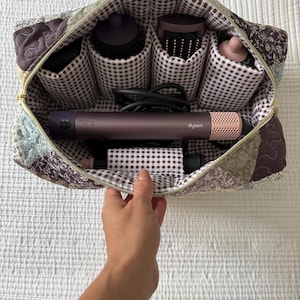 Könnte beinhalten: Eine gesteppte Aufbewahrungstasche mit braun-weiß kariertem Innenfutter und mehreren zylindrischen Fächern. Im Inneren liegt ein lilafarbenes und rosafarbenes Dyson-Haarstylinggerät horizontal, umgeben von verschiedenen Styling-Aufsätzen.