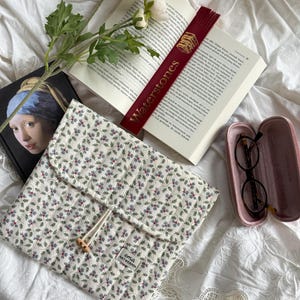 Könnte beinhalten: Eine Buchhülle mit floralem Muster und einem Knopfverschluss aus Holz, ein weinrotes Lesezeichen mit dem Text "Waterstones" und eine Brille in einem rosa Etui. Ein Buch und eine weiße Blume sind ebenfalls im Bild.