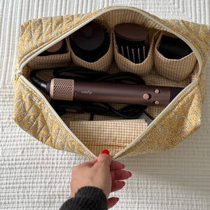 Op de afbeelding: Open gele, gewatteerde cosmetische tas met bloemenpatroon en ritssluiting, met een Dyson haarstylingtool en diverse borstelopzetstukken. De tas heeft een gingham voering.