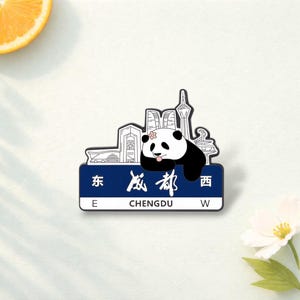 May include: A rectangular magnet featuring a panda bear resting above a blue sign with the word "Chengdu" in white. The magnet also includes a cityscape silhouette and the text "E" and "W".