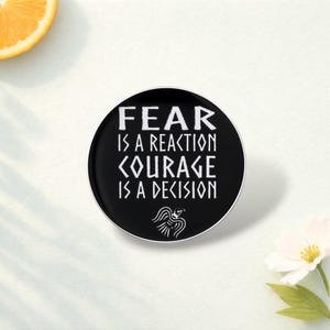 May include: A round, black pin with white text that reads "FEAR IS A REACTION COURAGE IS A DECISION." A small, white bird graphic is at the bottom. The pin is on a white surface with a slice of orange.