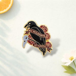 May include: A gold-toned enamel pin featuring a black raven perched in front of red roses. A banner across the bird displays the text NO MOURNERS NO FUNERALS in gold lettering. A pale blue plague doctor mask is positioned to the left of the raven.