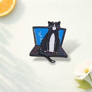 Könnte beinhalten: Eine schwarz-weiße Katzen-Emaille-Anstecknadel, die auf einer Laptop-Tastatur sitzt. Der Laptop-Bildschirm ist blau mit einem traurigen Smiley und dem Text :( Be. Die Tastatur hat einen regenbogenfarbenen Verlaufseffekt auf den Tasten.