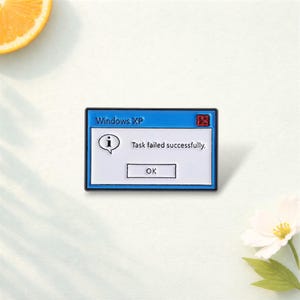 May include: A blue and white enamel pin in the shape of a Windows XP error message. The pin displays the text "Windows XP", "Task failed successfully.", and an "OK" button. An "i" icon is also visible.
