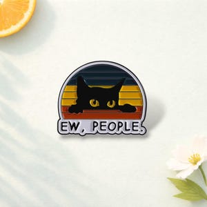 May include: A semi-circular enamel pin featuring a black cat silhouette with yellow eyes peering over a ledge. The background consists of horizontal stripes in dark blue, yellow, orange, and red. The white text below reads, "EW, PEOPLE."