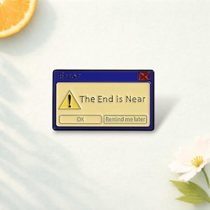 Op de afbeelding: Een rechthoekige emaille speld in de vorm van een blauw computermeldingvenster met een crèmekleurig midden. Het toont een gele waarschuwingsdriehoek met een uitroepteken en de tekst "Error", "The End is Near", "OK" en "Remind me later".