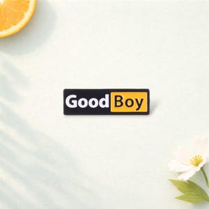 May include: A rectangular pin with the words "Good Boy" in white text on a black background, with the word "Boy" in a yellow box. The pin is on a white surface, with a slice of orange and a white flower in the background.