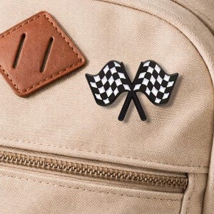 Puede incluir: Una mochila beige con un parche de cuero marrón y un pin de bandera a cuadros en blanco y negro. El pin presenta dos banderas cruzadas, cada una con un patrón de cuadros en blanco y negro. La mochila tiene una cremallera y un parche de cuero marrón.