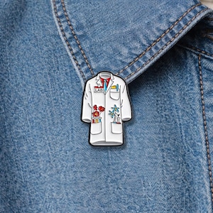 May include: A white lab coat enamel pin with black trim, featuring a red tie, blue tie, and pockets filled with flowers and lab equipment. The pin is set against a blue denim jacket.