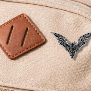 Könnte beinhalten: Ein silberfarbener Fledermaus-Ansteckpin mit detaillierten Flügeln und Körpermerkmalen, auf einem beigefarbenen Rucksack. Neben dem Pin befindet sich ein brauner Lederaufnäher mit zwei Schlitzen.