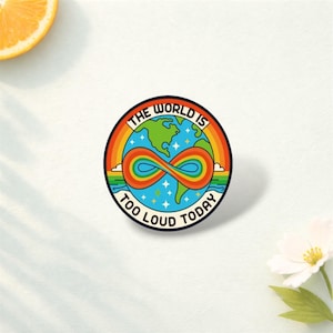 May include: A circular sticker with a vibrant design. It depicts a globe in green and blue, with a rainbow infinity symbol. The text "THE WORLD IS TOO LOUD TODAY" is printed around the edge of the sticker.