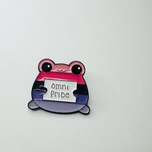 Könnte beinhalten: Eine Metall-Emaille-Anstecknadel in Form eines Frosches mit großen schwarzen Augen. Der Frosch hat horizontale Streifen in den Farben Pink, Schwarz und Lila. Er hält ein weißes rechteckiges Schild mit dem schwarzen Text "omni pride" in der Mitte.