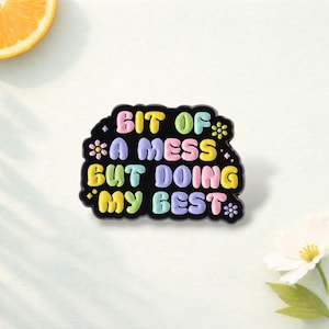 Pode incluir: Um broche de esmalte preto com uma borda ondulada e irregular, apresentando o texto colorido "BIT OF A MESS BUT DOING MY BEST" em letras arredondadas. O design inclui pequenos detalhes de flores e estrelas em tons pastel amarelo, rosa e azul.