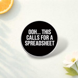 May include: A black circular phone grip with the white text "OOH... THIS CALLS FOR A SPREADSHEET". It has a silver edge and is on a white surface, with a slice of orange and a white flower in the background.