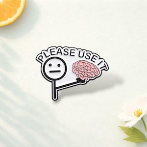 May include: A white enamel pin featuring a black stick figure with a neutral expression, holding a pink brain illustration. The text "PLEASE USE IT" is printed in black capital letters along the top edge of the pin's white background.
