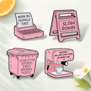 May include: Four pink enamel pins with black outlines. The pins include a typewriter with the text "WORK ON YOURSELF FIRST", a road sign that says "SLOW DOWN", a trash can with "UNSOLICITED OPINIONS FROM RANDOM PEOPLE", and an espresso machine with the text "you can't pour from an empty cup".