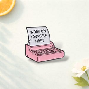 May include: A pink typewriter enamel pin with the text "WORK ON YOURSELF FIRST" on a white paper. The pin has a black outline and is set against a white background with a slice of orange and a white flower.