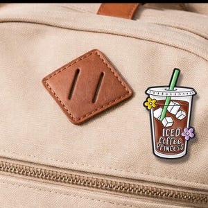 May include: A tan backpack with a brown leather patch and a colorful enamel pin. The pin features an iced coffee cup with the text "Iced Coffee Princess" and floral accents. The pin has a green straw and ice cubes.