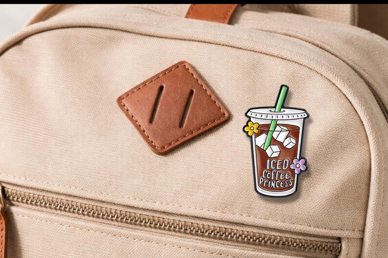 May include: A tan backpack with a brown leather patch and a colorful enamel pin. The pin features an iced coffee cup with the text "Iced Coffee Princess" and floral accents. The pin has a green straw and ice cubes.