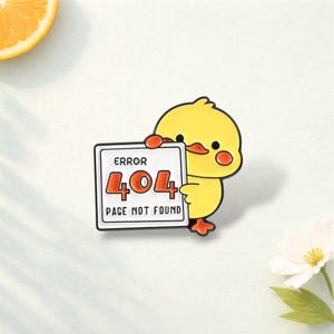 Könnte beinhalten: Ein gelber Küken-Anstecker, der ein Schild mit der Aufschrift "ERROR 404 PAGE NOT FOUND" hält. Das Küken hat orangefarbene Wangen und Füße. Das Schild hat einen weißen Hintergrund mit schwarzen Rändern und orangefarbenem Text. Ein lustiges Accessoire.