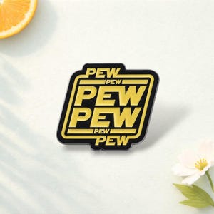 May include: A black and yellow enamel pin with the word "PEW" repeated three times in a rectangular design. The pin has a black border and a yellow interior. The pin is on a white surface with a slice of orange and a flower.