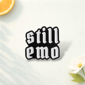 Può includere: Spilla in smalto bianco e nero con la scritta "still emo" in un carattere audace e stilizzato. La spilla è posta su uno sfondo bianco, con una fetta d'arancia e un fiore bianco.