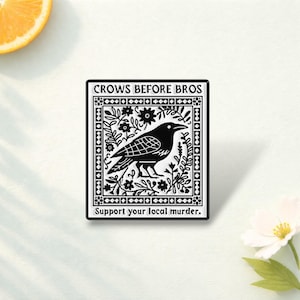 May include: A rectangular black and white enamel pin featuring a central illustration of a crow surrounded by floral patterns. The text "CROWS BEFORE BROS" is printed at the top, and "Support your local murder." is printed at the bottom.