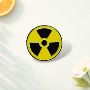 May include: A circular enamel pin featuring a bright yellow background with a black radiation hazard symbol in the center. The design consists of three black fan-shaped segments arranged around a central black circle, all enclosed by a thin black border.